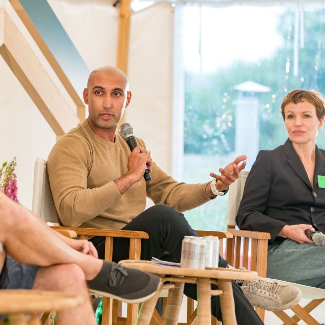 A man holding a microphone speaks while seated on a panel with two other people during an outdoor event.